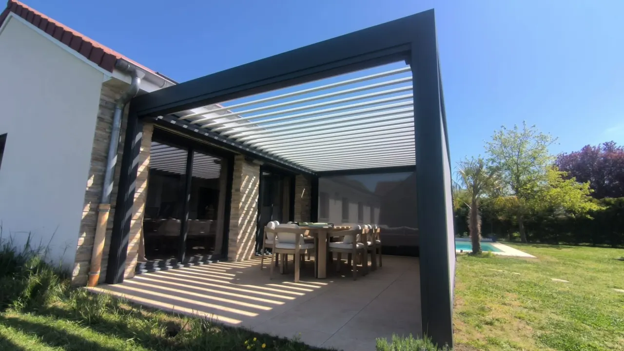 Pergola bioclimatique en aluminium anthracite adossée à une maison, lames orientables, store zip et table de repas sur la terrasse