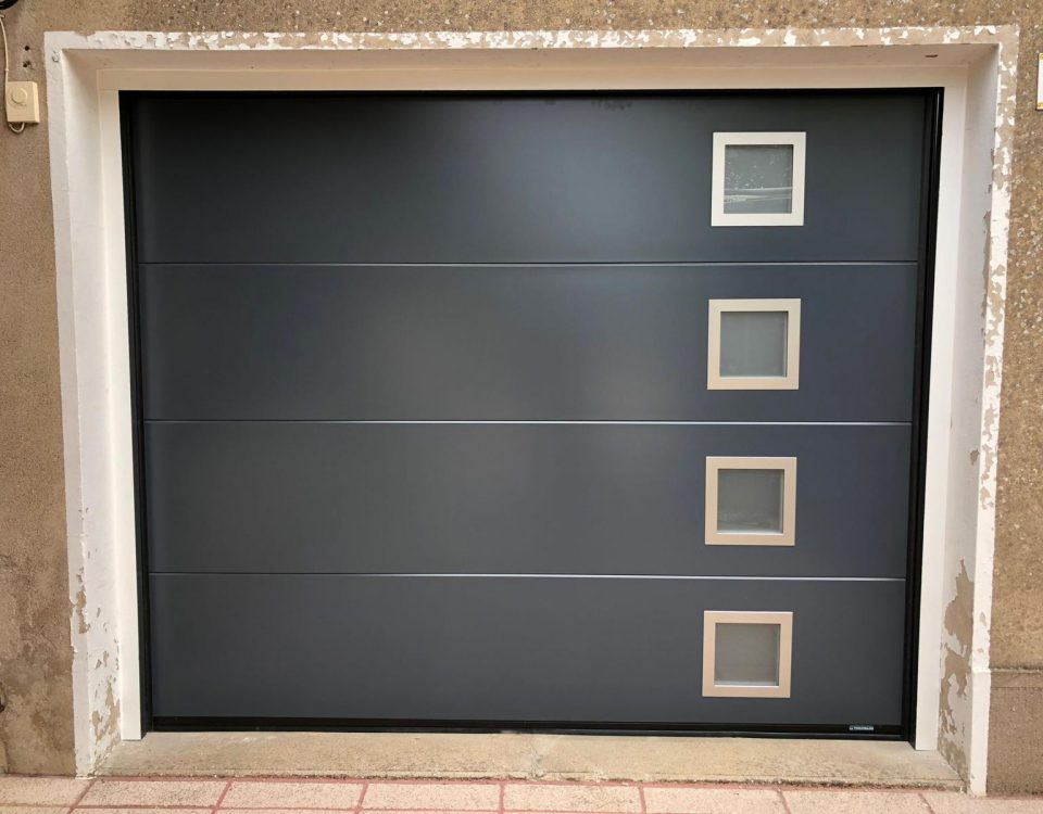 Porte de garage sectionnelle en aluminium laquée gris anthracite avec épaisseur de 42mm isolante avec manoeuvre motorisée et design lisse avec hublots