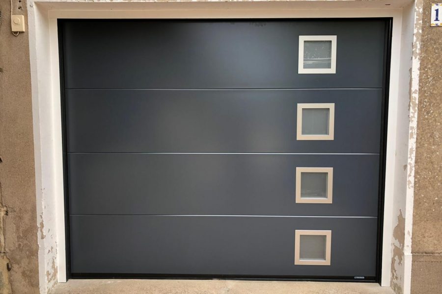 Porte de garage sectionnelle en aluminium laquée gris anthracite avec épaisseur de 42mm isolante avec manoeuvre motorisée et design lisse avec hublots